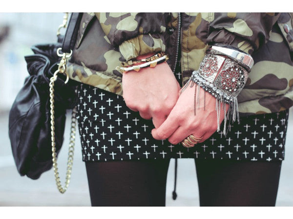 A person in a camo jacket and black skirt with white crosses is zipping up a camo jacket. They wear multiple bracelets and hold a black bag with a gold chain.