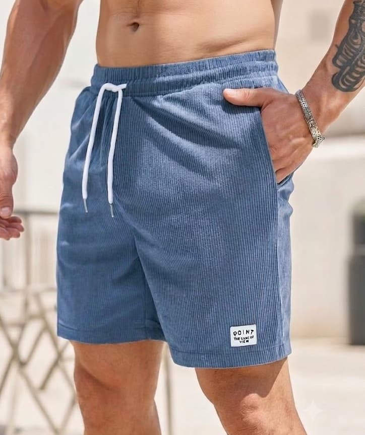 A close-up of a man wearing blue corduroy drawstring shorts with a white patch near the hem, set against a blurred outdoor background.