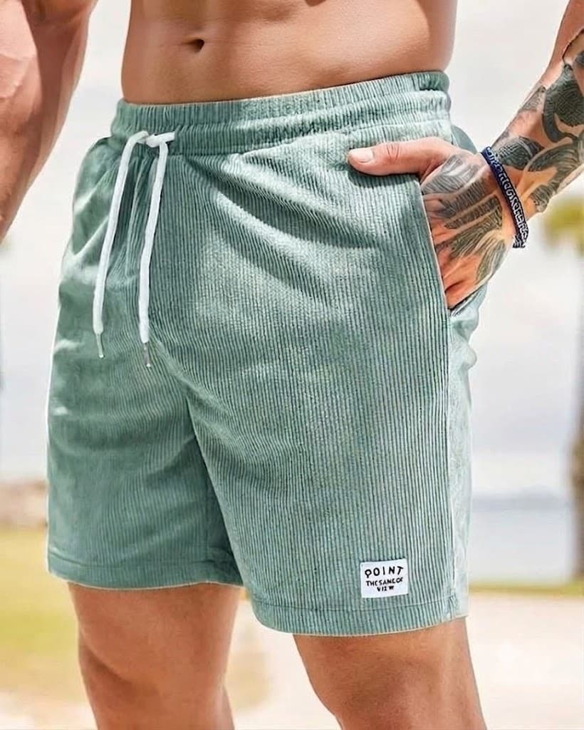 A close-up of a person wearing sage green corduroy shorts with a white drawstring and a small white patch near the hem. The person's hand is in the pocket, and the background is a softly blurred outdoor setting.