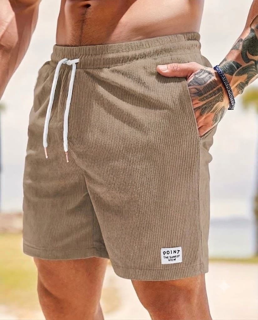 A close-up of a man wearing taupe corduroy shorts with a white drawstring and a small white patch near the hem. The background is a blurred beach setting.