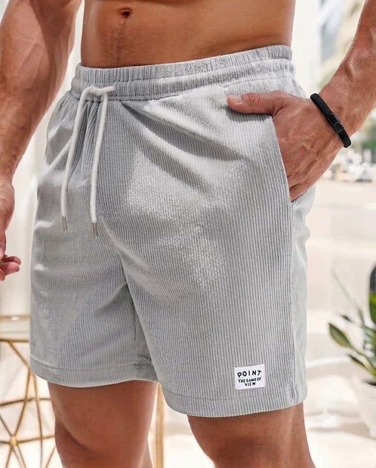 A close-up of a man wearing light gray ribbed drawstring shorts with a small white label near the hem, shown in a bright indoor setting.