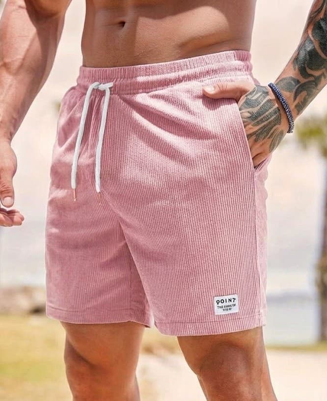 A close-up of a man wearing pink ribbed corduroy shorts with a white drawstring and a small white patch near the hem, set against a blurred beach background.