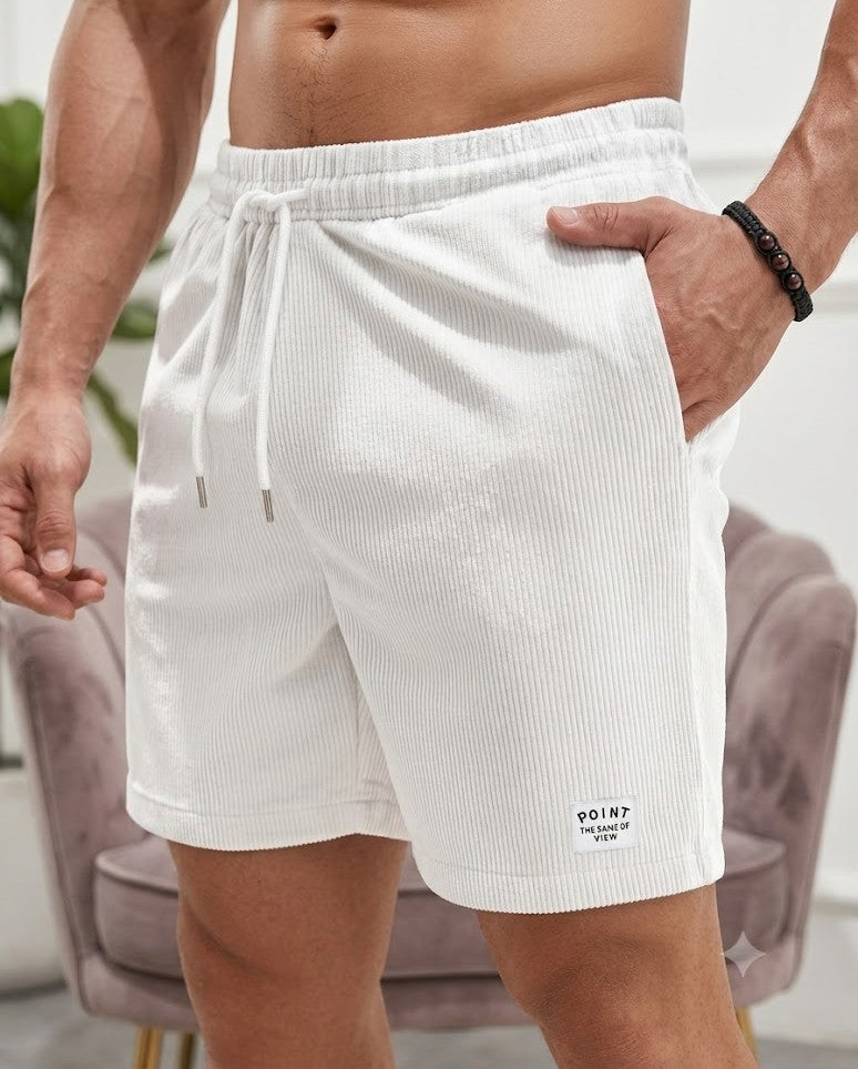 A close-up of a man wearing white ribbed drawstring shorts, with his hand in his pocket. The shorts feature a small white patch on the lower left leg