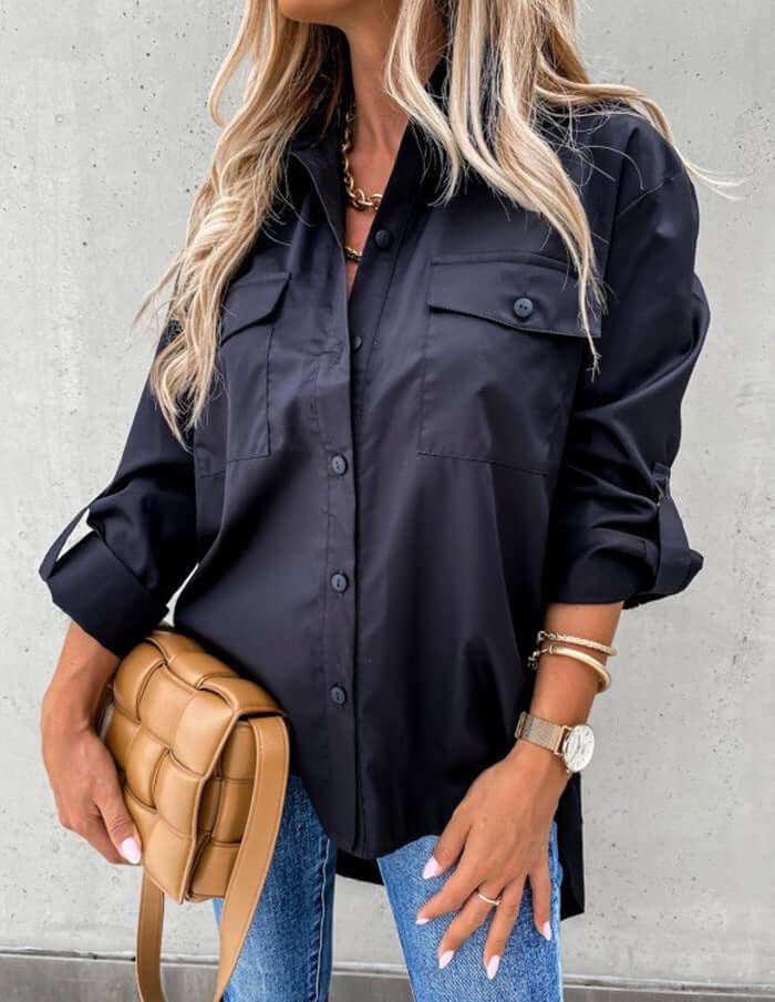 Person wearing a black button-up shirt and blue jeans, holding a tan handbag against a gray background